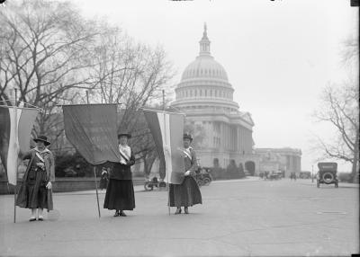 three women in black and white photo with signs and long dresses and hats standing in front of capitol