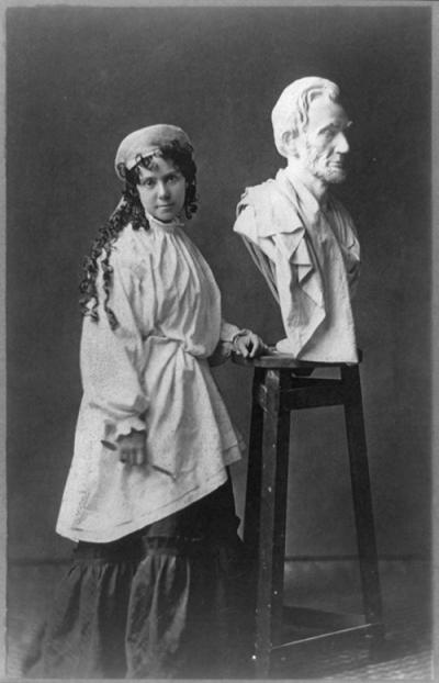 black and white photo of a girl with long curly hair and a white dress standing in front of a white marble bust of abraham lincoln