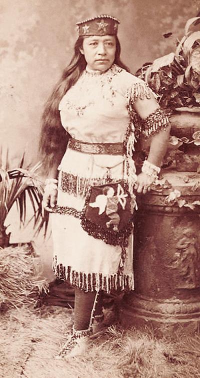 black and white photo of a woman standing in traditional native american clothing in grass near a sculpture