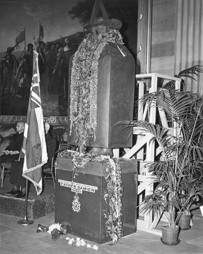 large black and white photo of a statue that has a name at the bottom stand and is sitting in front of a large painting in the background