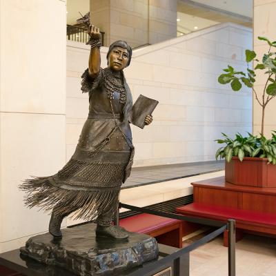 bronze statue of a woman walking forward and her dress blowing in the wind. she's holding a book and pointing ahead with her other hand