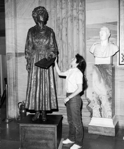 a black and white photo of a woman in jeans touching and staring the bronze statue of a woman in a dress holding a piece of paper. to her right is another bust