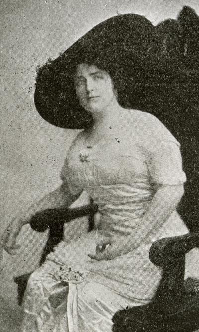 black and white photo of a woman sitting in a chair with a dress and large black hat on