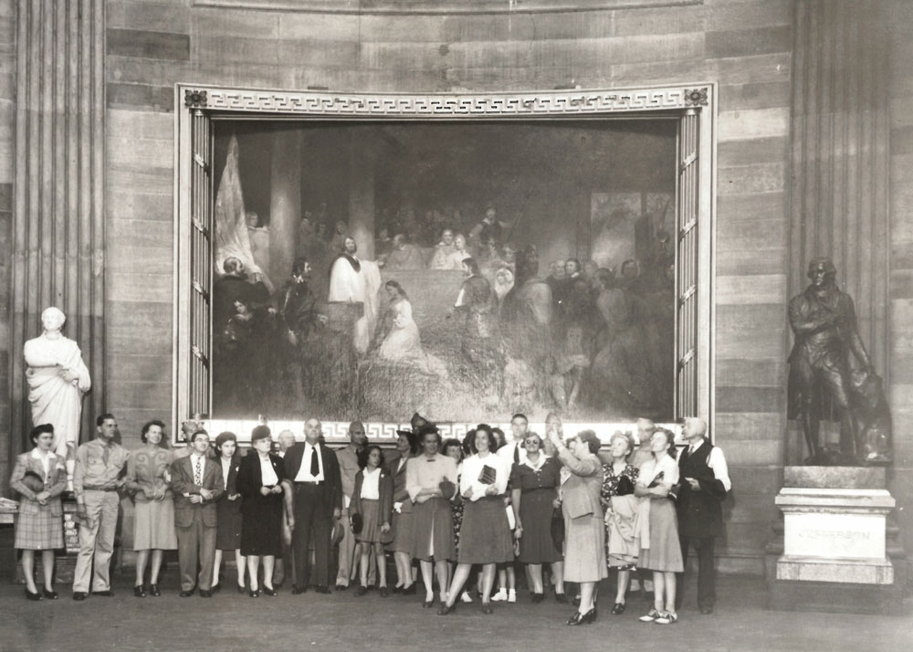 Black and white photo of a large group in front of a painting depicting the baptism of Pocahontas.