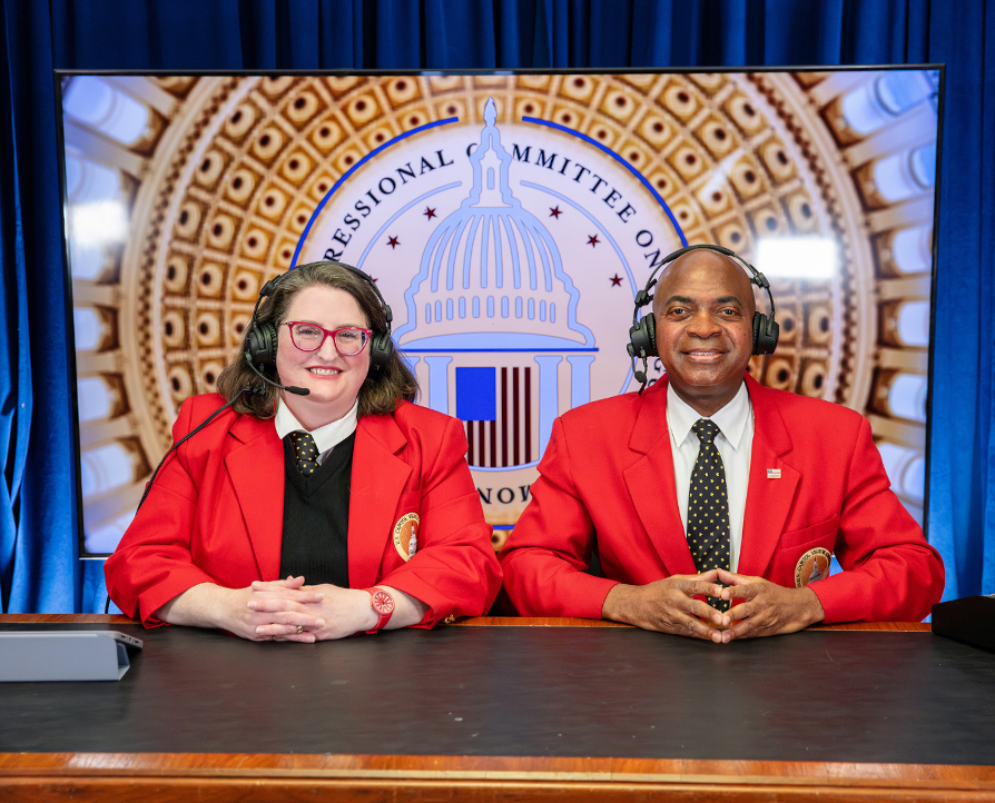 A woman and man wearing headsets and sitting at a desk smile in front of a backdrop