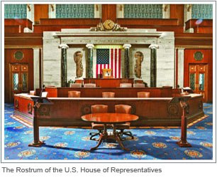 The rostrum of the house of representatives with burgundy wood, white marble background, and seats in front of it for a speaker 