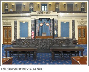 The senate rostrum with black accents behind the podium and a blue rug and marble corners surrounding the speakers podium and rows of chairs in front of it 