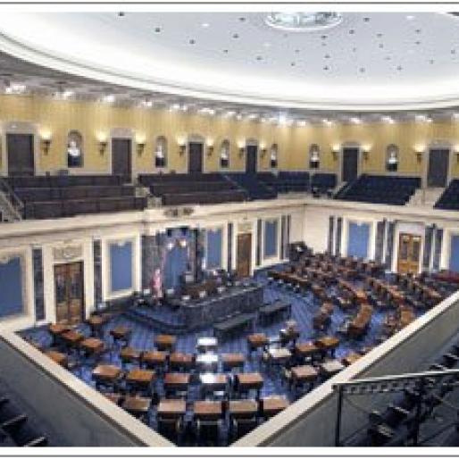 Senate chamber with a circular light in the ceiling shining above rows of chairs all facing a speakers podium in a half-circle shape