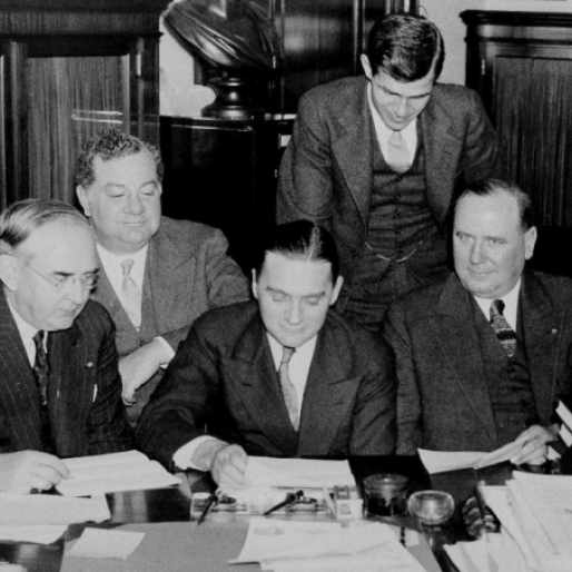 group of five congresspeople wearing suits and ties; three are seated at a table with documents laid in front of them while two additional people observe from behind them