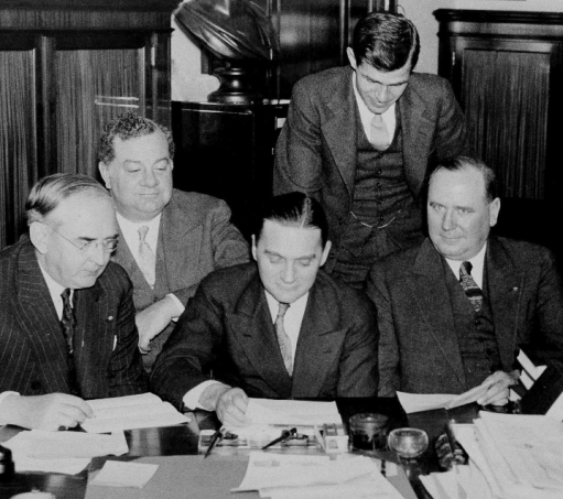 group of five congresspeople wearing suits and ties; three are seated at a table with documents laid in front of them while two additional people observe from behind them