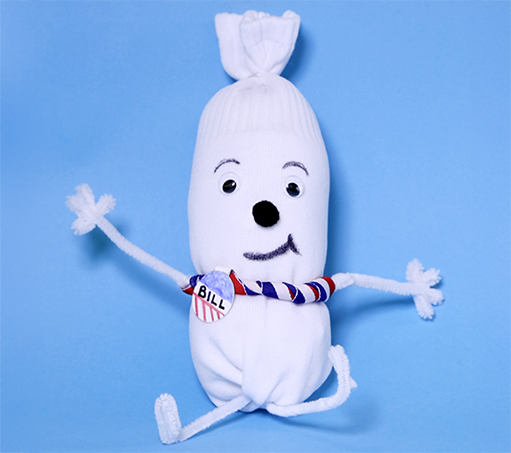 handmade toy on a light blue background; the toy is a stuffed white sock tied off at the top and adorned with googly eyes, black pom-pom nose, Sharpie-drawn eyebrows and smile, pipe cleaner arms and legs, and a red, white, and blue striped ribbon with the word “BILL” tied around it