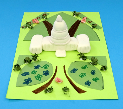 handmade model of the U.S. Capitol crafted from white clay; the model is sitting on top of a piece of green construction paper adorned with slightly darker green construction paper pieces representing grass and crumpled paper strips to represent bushes on a bright blue background
