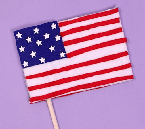 handmade American flag on a light purple background; the flag is crafted from red, white and blue felt attached to a miniature wooden stick 