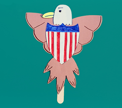 handmade eagle and Union Shield on a teal background; the eagle is crafted from white and brown construction paper and a popsicle stick and its wings are open around either side of the shield