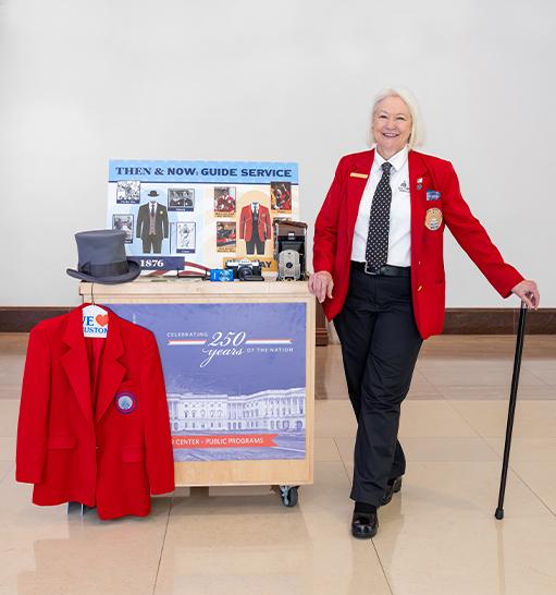Visitor Guide wearing a red blazer stands next to the Then and Now education cart