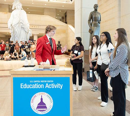Visitor Guide in a red blazer speaks to a group of four visitors at a blue Education Activity cart in front of the Statue of Freedom