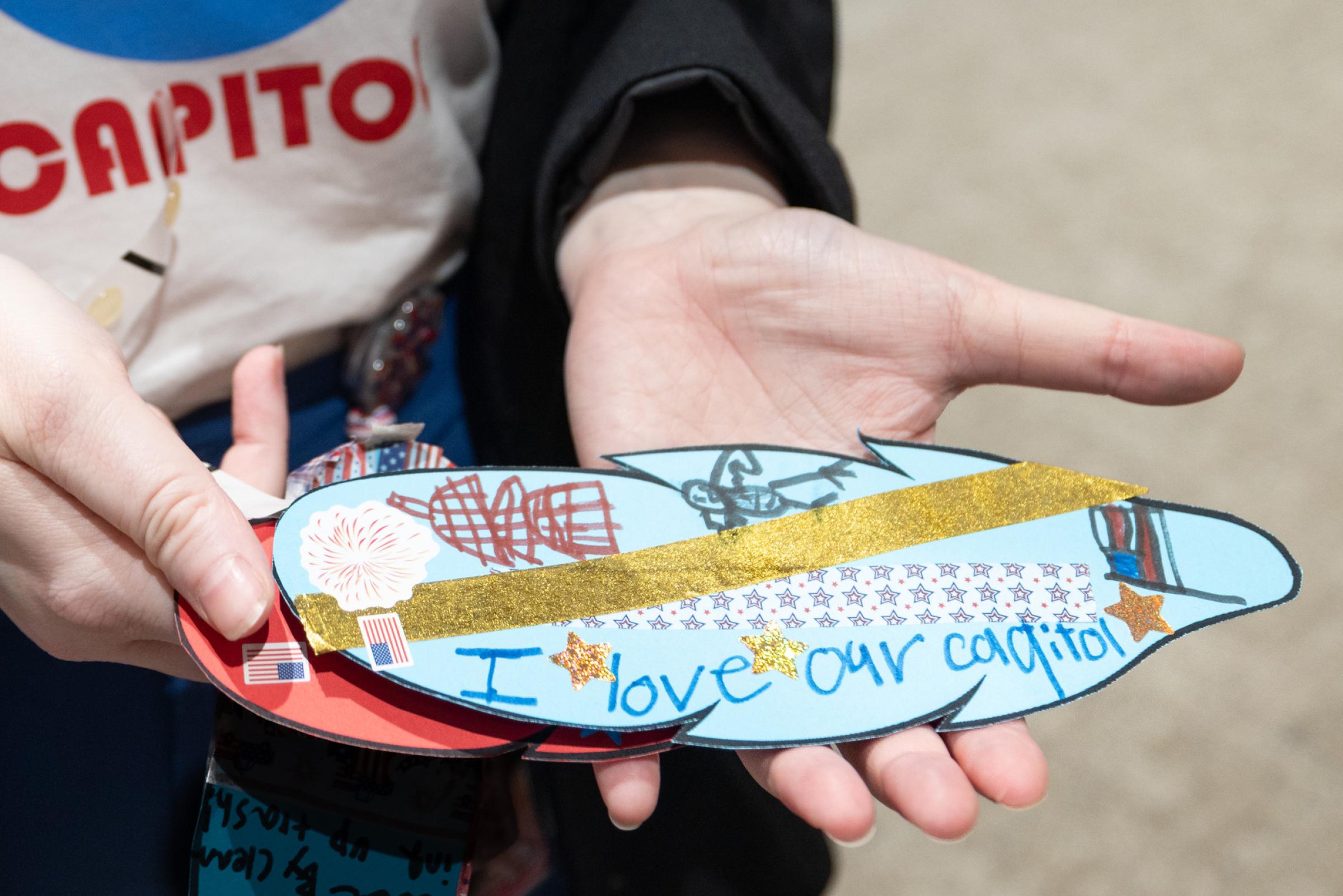 two hands holding a paper feather decorated with the words I love our Capitol