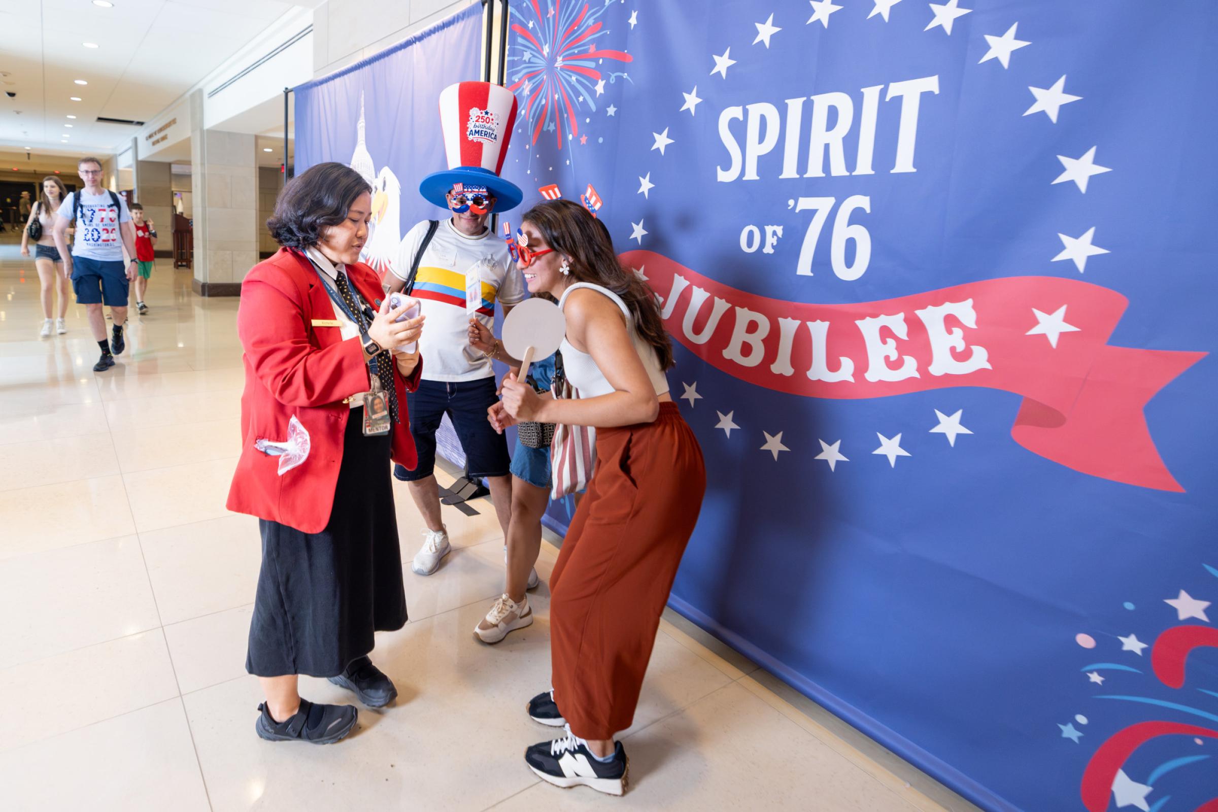 Visitor Guide wearing a red blazer assists visitors in front of a photobooth backdrop