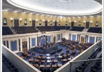 Senate chamber with a circular light in the ceiling shining above rows of chairs all facing a speakers podium in a half-circle shape