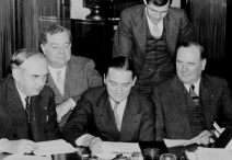 group of five congresspeople wearing suits and ties; three are seated at a table with documents laid in front of them while two additional people observe from behind them