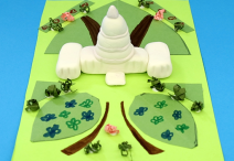 handmade model of the U.S. Capitol crafted from white clay; the model is sitting on top of a piece of green construction paper adorned with slightly darker green construction paper pieces representing grass and crumpled paper strips to represent bushes on a bright blue background