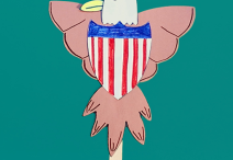 handmade eagle and Union Shield on a teal background; the eagle is crafted from white and brown construction paper and a popsicle stick and its wings are open around either side of the shield
