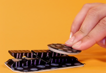 handmade telegraph key on an orange background; the device is crafted from a black wooden tag and black clothespin that have been decorated with metallic stripes and circles, and  three fingers push the device’s button from the right side