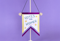 handmade “VOTES FOR WOMEN” banner on a purple background; the miniature banner is crafted from construction paper adorned with purple and yellow ribbon and mounted to a popsicle stick hanging from a black string
