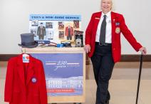Visitor Guide wearing a red blazer stands next to the Then and Now education cart