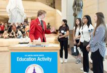 Visitor Guide in a red blazer speaks to a group of four visitors at a blue Education Activity cart in front of the Statue of Freedom