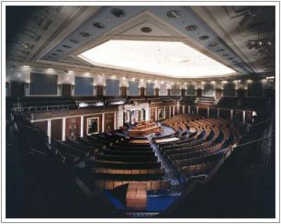 the house chamber of the capitol, dark room with light coming from the ceiling and lots of rows of chairs facing a speakers podium