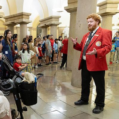 Visitor Guide wearing a red blazer gesturing with both hands to a large group of visitors in the Crypt