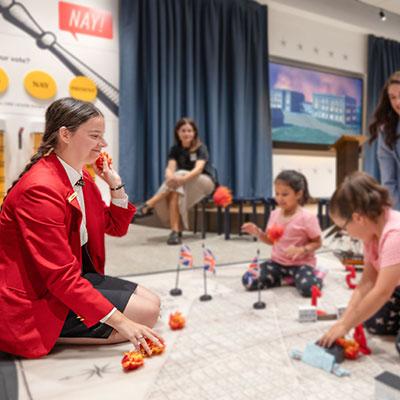 Visitor Guide wearing a red blazer kneels on the floor while explaining something to two children