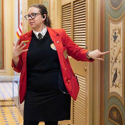 Visitor Guide in a red blazer pointing at a small painting on a wall