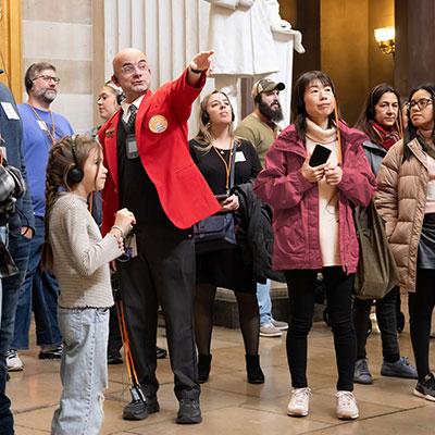 Visitor Guide wearing a red blazer points out something across the room to a group of visitors
