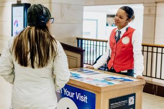 A visitor assistant in a red vest greets a guest.